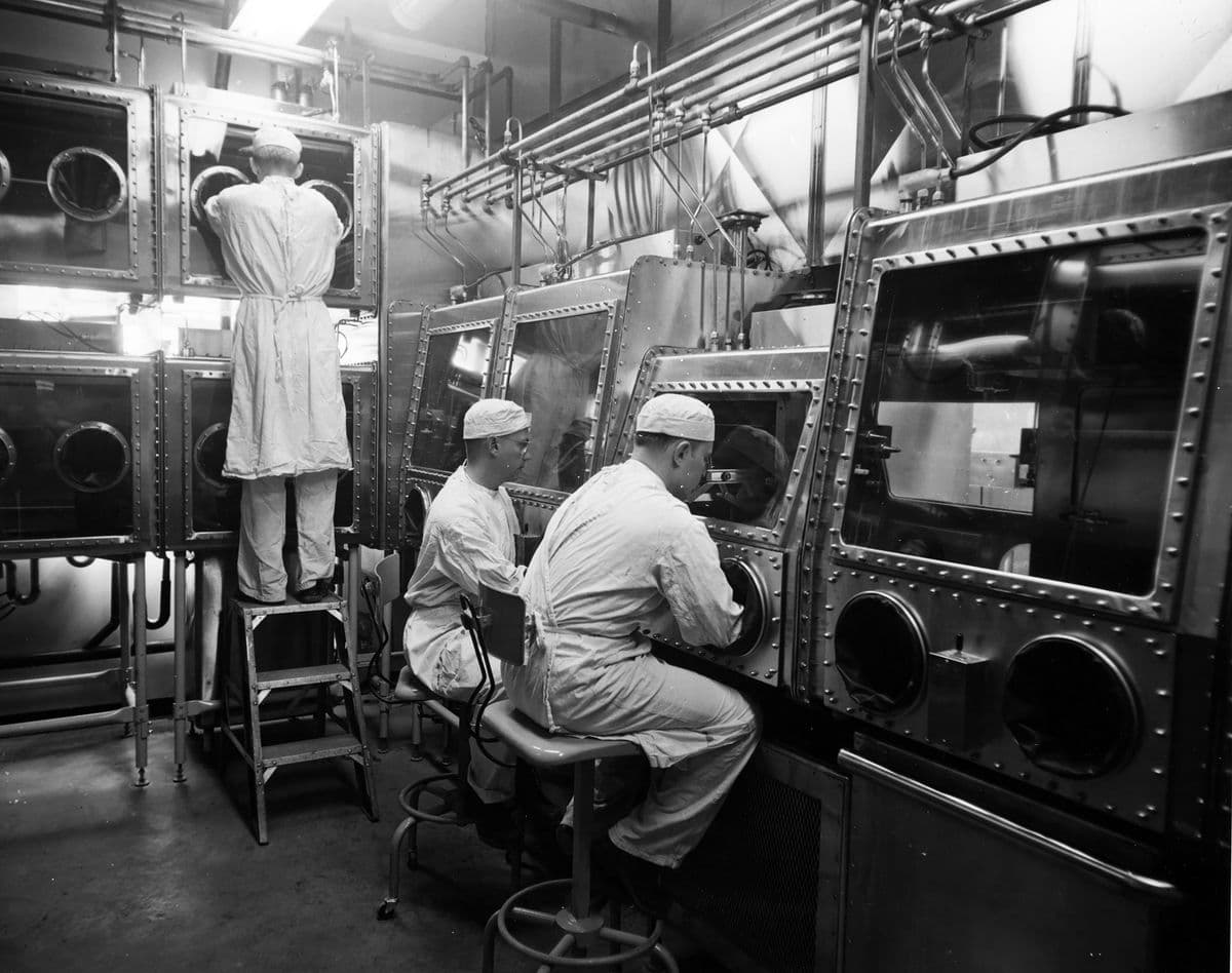 画像は実験室で働く3人の作業員で、全員が防護服を着用し、手袋が内蔵された密閉区画で材料を扱っている。