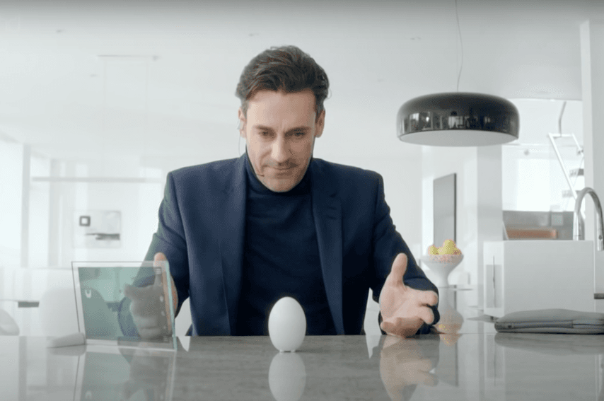 モダンなキッチンで、スーツ姿の男性が磨き上げられた表面にもたれかかり、水平に置かれた卵型の電化製品を興味深そうに見つめている。好奇心や驚きの雰囲気が感じられる。