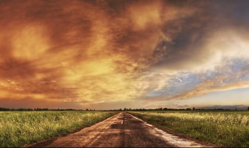 画像には、平野を横切るまっすぐな道路が描かれており、左側にはオレンジや黄色の色調を映し出す雲がドラマチックに、右側には澄み切った明るい空が広がる緑の野原が広がっている。