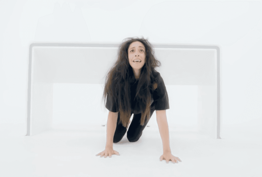 白い部屋で床に手をついてひざまずく女性の表情は、恐怖と苦しみを表している。